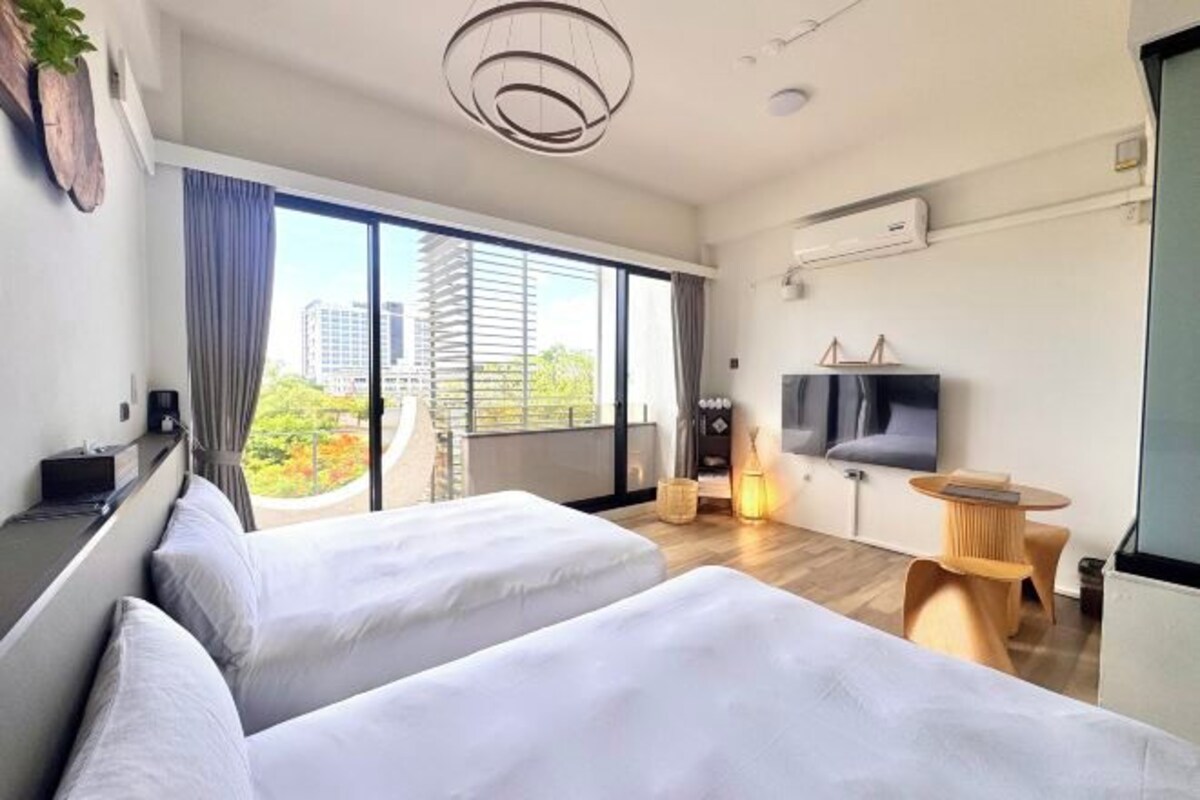 A modern hotel room with two beds, a TV, a small table, and a large window offering a city view.