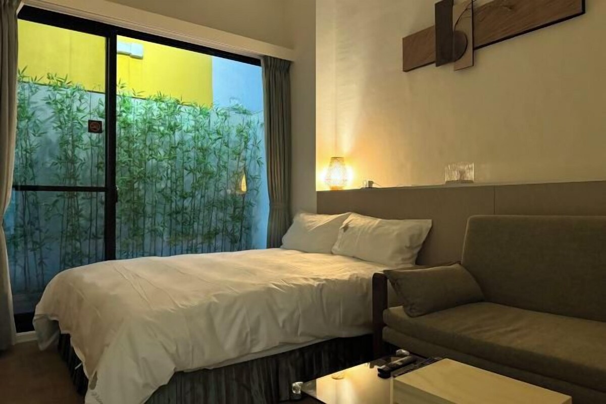 A hotel room with a bed, a sofa, a bedside table, and a window with a view of bamboo outside.