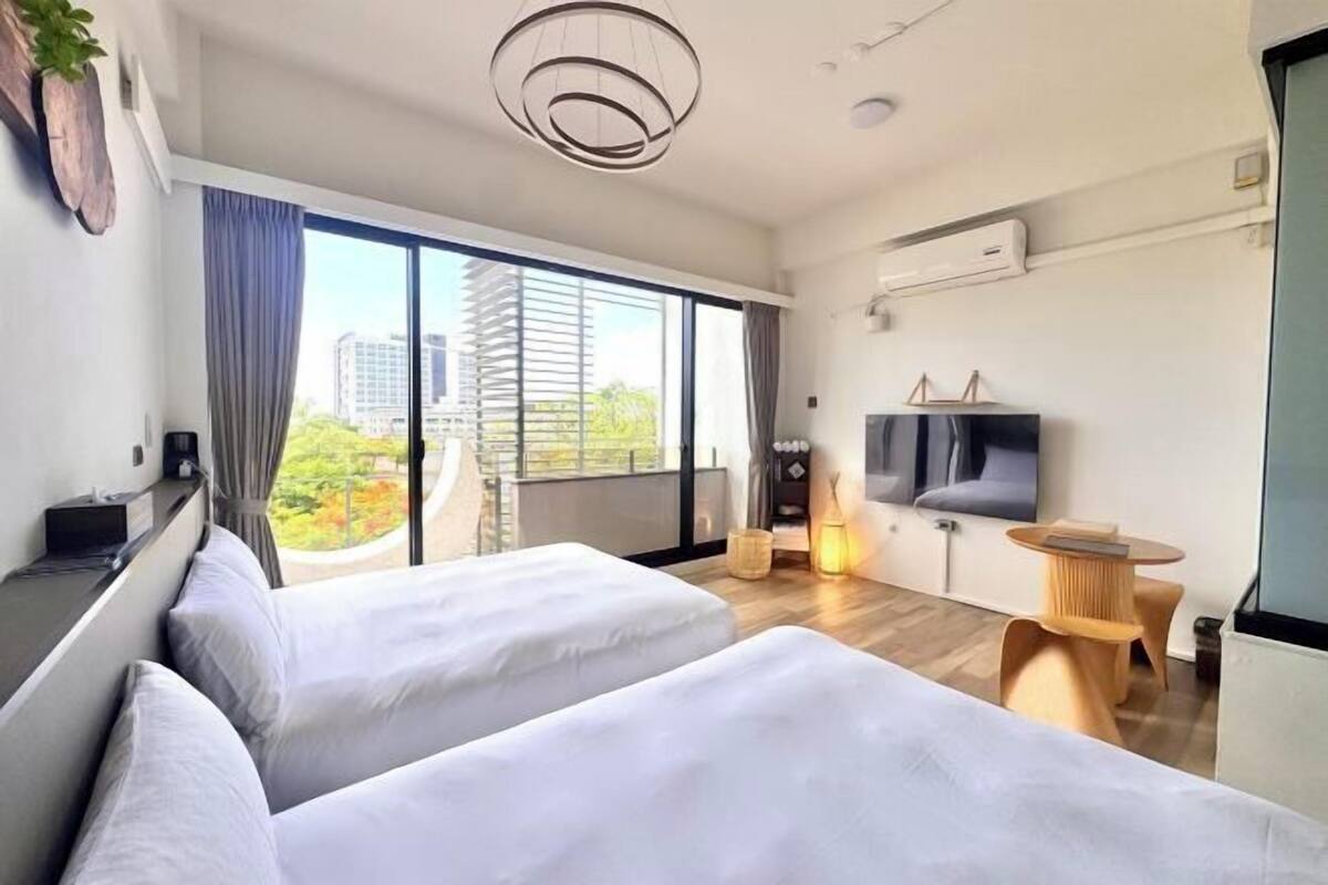 A modern hotel room with two beds, a TV, a small table, and a large window offering a city view.