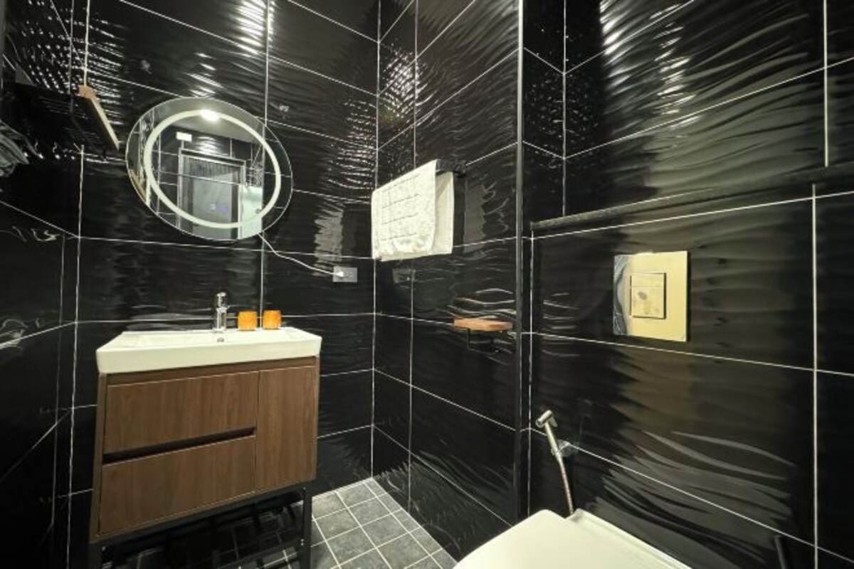 A modern bathroom with black tiled walls, a wooden vanity, a round mirror, and a white sink.