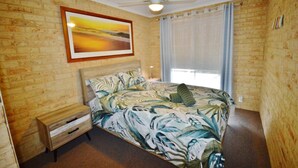 Un dormitorio con pared de ladrillo, una cama con colcha estampada, una mesita de noche, un ventilador de techo y una ventana con cortinas.