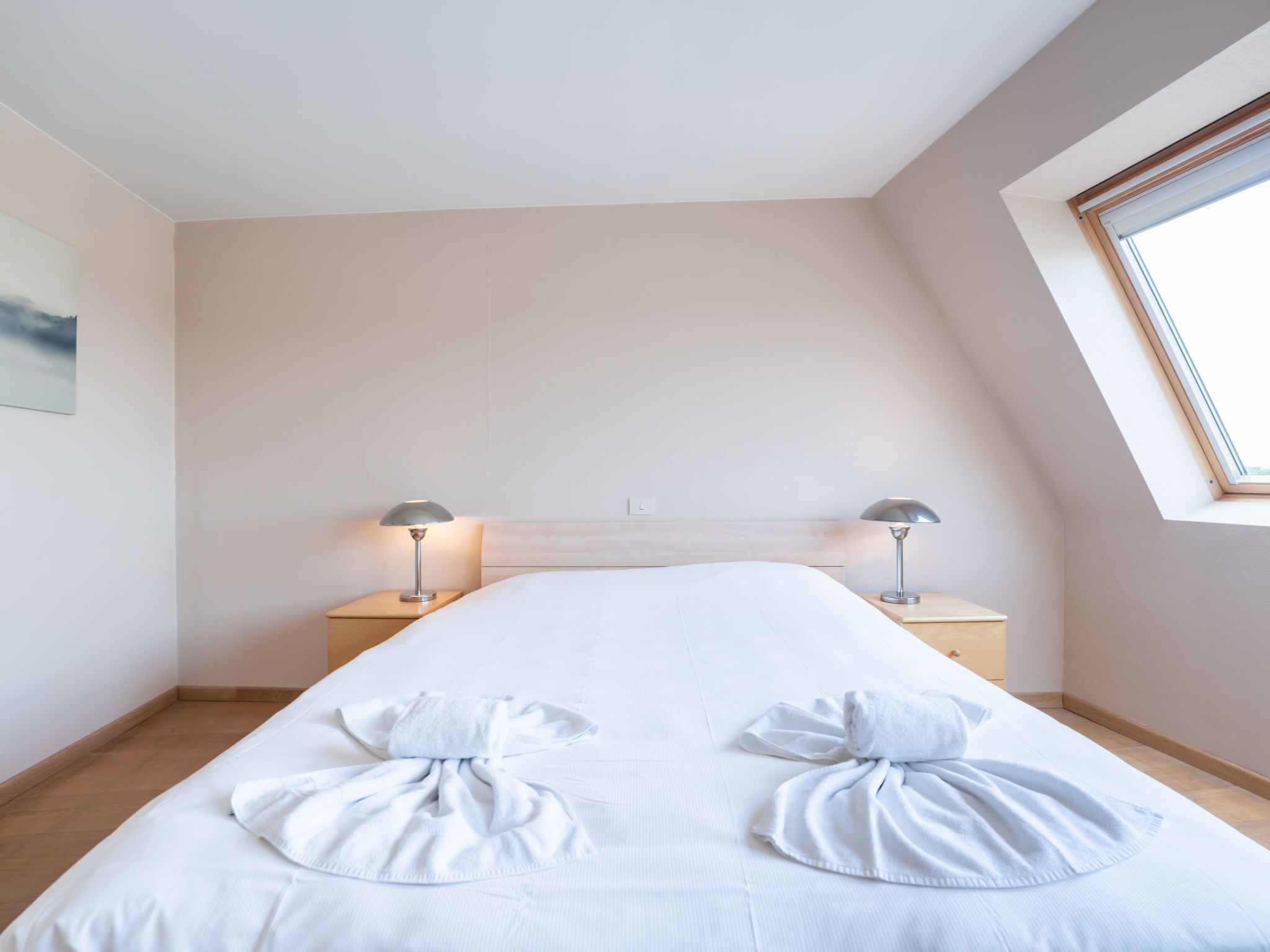 A modern hotel room with a large bed, two bedside tables with lamps, a skylight, and a painting on the wall.