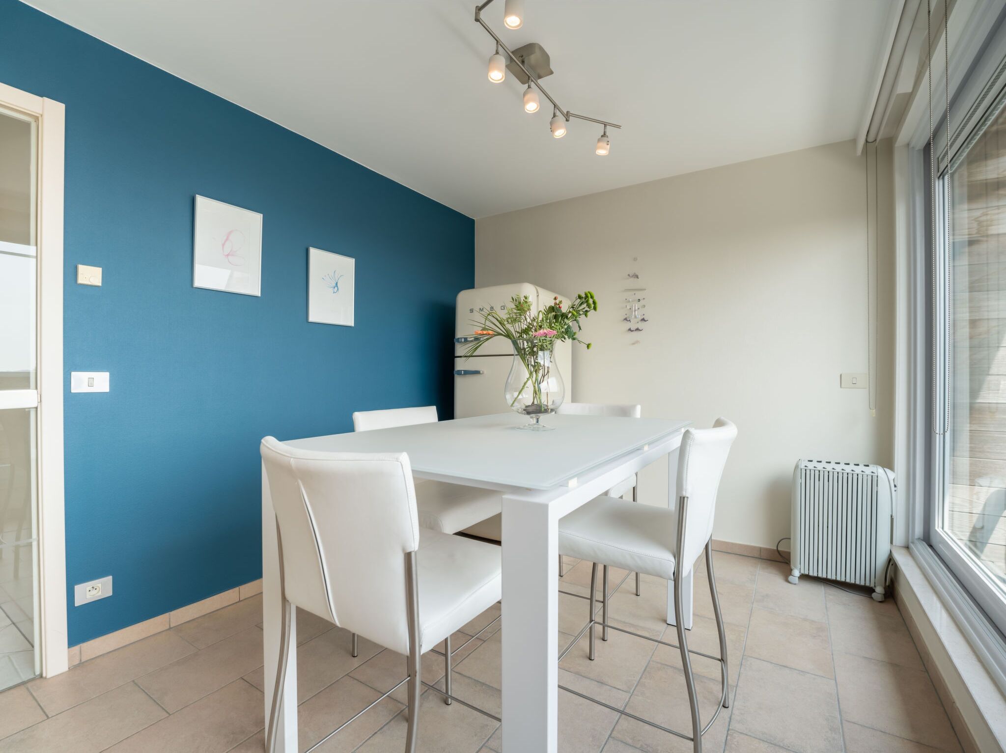 Eine moderne Küche mit einem weißen Tisch und Stühlen, einer blauen Akzentwand und einem Fenster mit Jalousien.