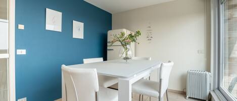 Eine moderne Küche mit einem weißen Tisch und Stühlen, einer blauen Akzentwand und einem Fenster mit Jalousien.