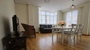 A spacious living room with a wooden floor, a dining table with chairs, a television, a sofa, and a large window with curtains.