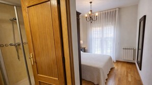 A hotel room with a wooden door, a shower, a bed with a white bedspread, a chandelier, and a radiator.