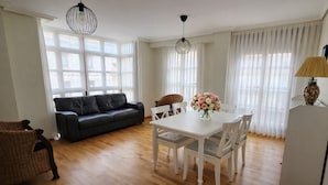 A spacious living area with a black leather sofa, a white dining table with chairs, and a wicker chair. There are two pendant lights above the table and a lamp on a stand to the right.