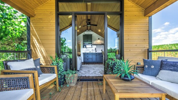 Una terraza de madera con un sofá, una mesa y una cocina visible a través de una puerta abierta.