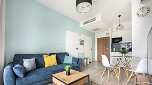 A modern living room with a blue sofa, a wooden coffee table, and a dining area with white chairs and a small table. - Prymasa Tysiąclecia Comfort Apartment (Warszawa)