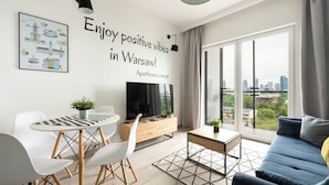 A modern living room with a TV, a dining table with chairs, a sofa, and a large window with a city view. - Prymasa Tysiąclecia Comfort Apartment (Warszawa)