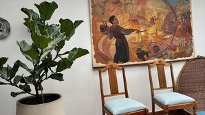 A room with a large potted plant, two wooden chairs with cushions, a colorful abstract painting on the wall, and a view through a skylight. - BLAU HOTEL CAFE (Sopó)