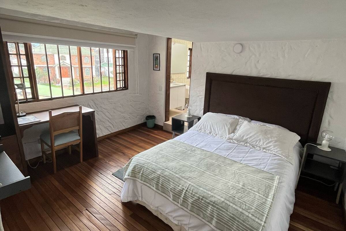 Ein Schlafzimmer mit einem großen Bett, einem Schreibtisch und einem Stuhl. Ein Fenster bietet Blick auf ein Gebäude.