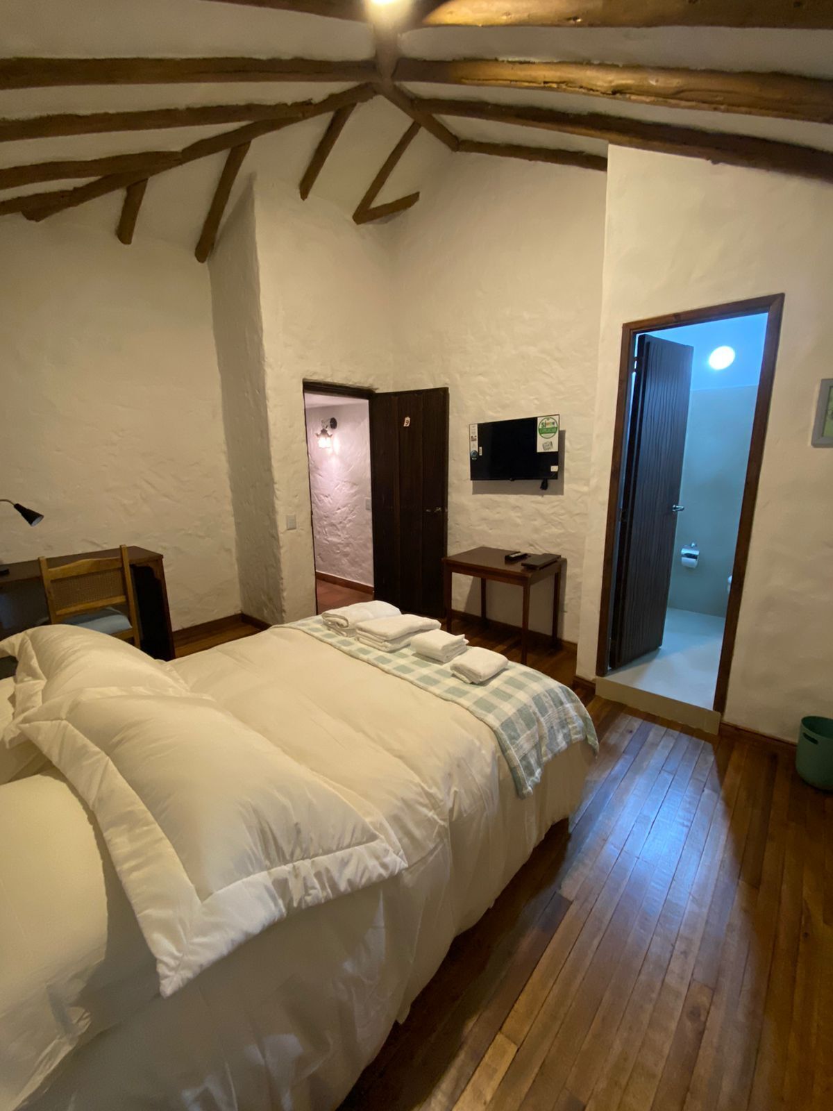 A bedroom with a large bed, wooden beams on the ceiling, a small table, and a TV mounted on the wall.