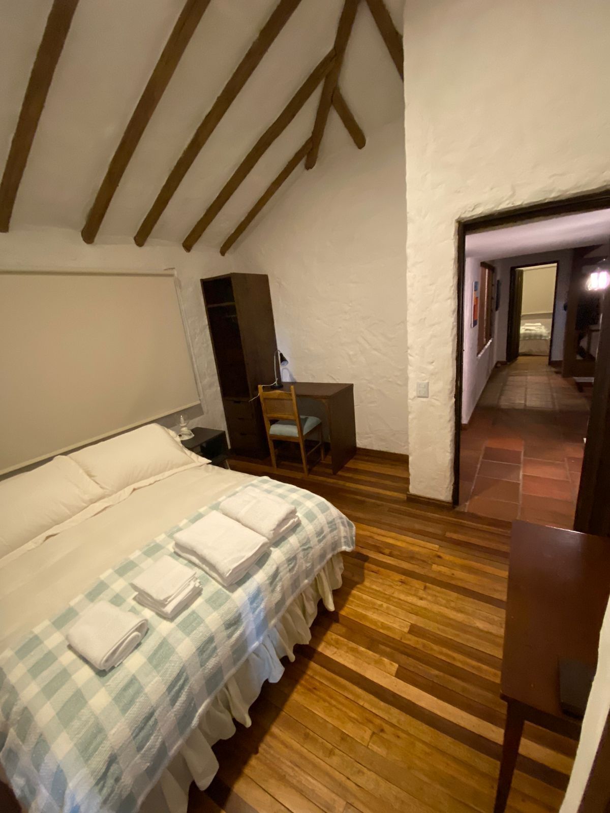 Un dormitorio con suelo de madera, una cama con colcha a cuadros, un escritorio con silla y techo inclinado con vigas de madera.