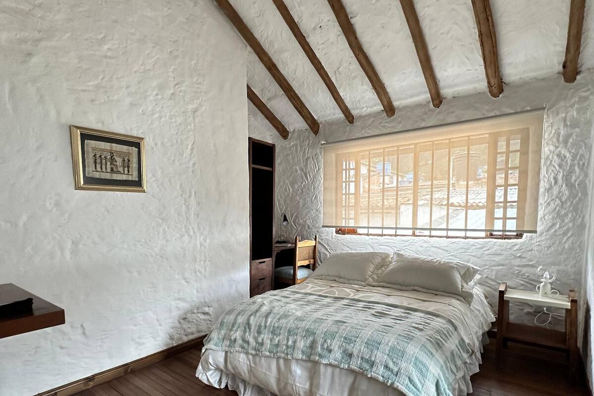 Ein Schlafzimmer mit Holzboden, einem Bett mit gemustertem Überwurf, einem Nachttisch mit Lampe und einem Fenster mit Gitterstäben.