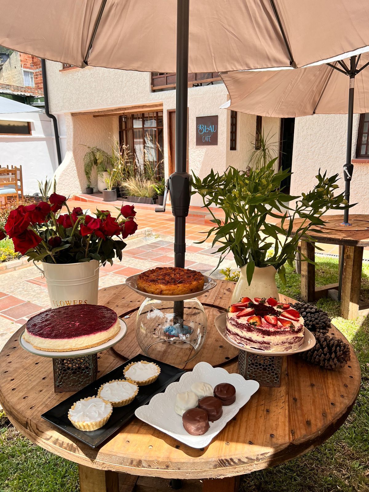 Una mesa de madera redonda con diversos postres y flores, ubicada al aire libre bajo un paraguas grande.