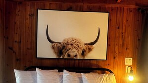 A wooden cabin interior with a bed, white pillows, a headboard, a wall-mounted artwork of a bull's head, and a nightstand with a lamp. - Secluded Nature Escape, Pet Friendly with WiFi in Three Rivers (Three Rivers)