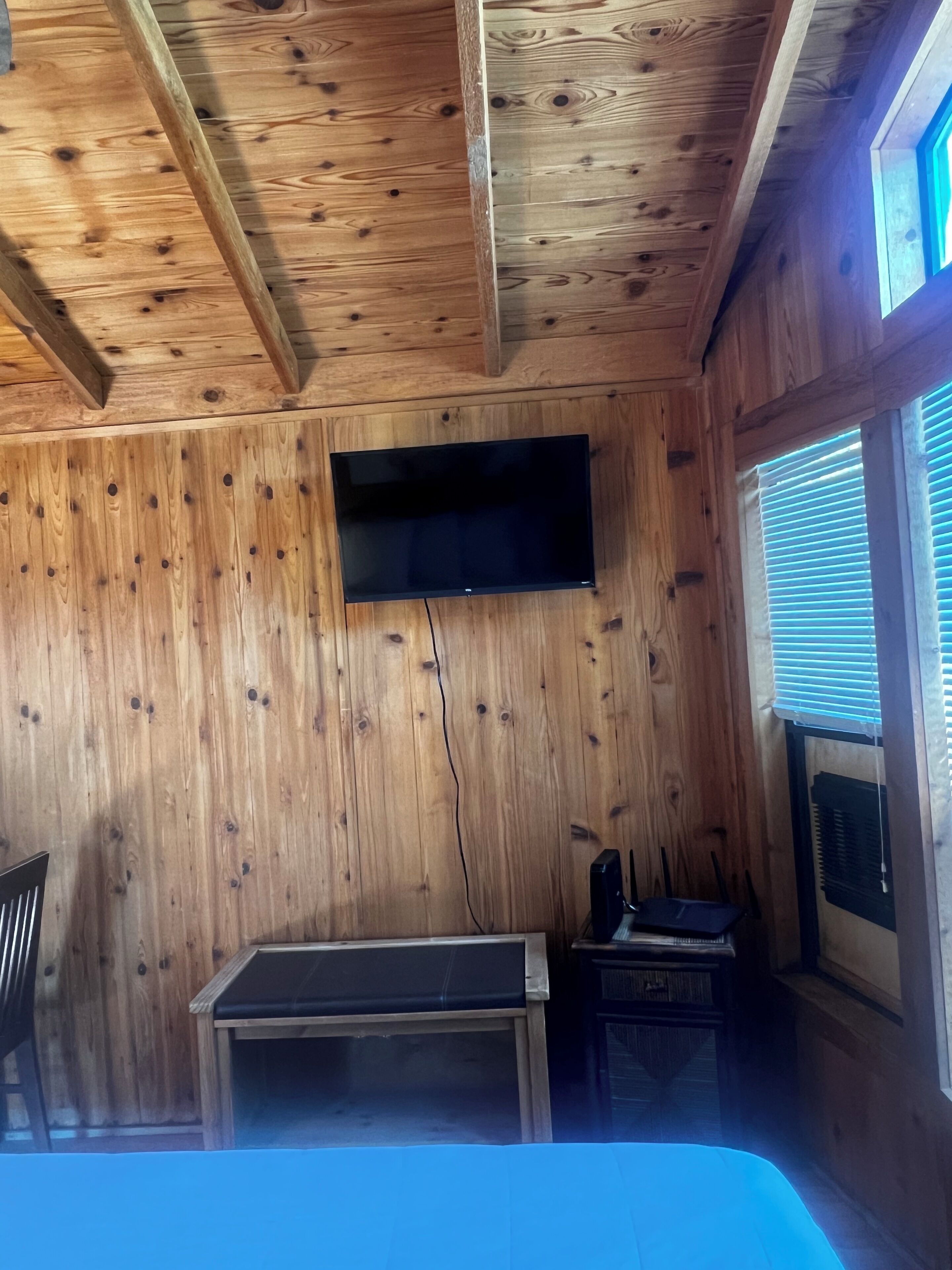 Una habitación con paredes de madera, un televisor de pantalla plana montado en la pared, un escritorio con una silla y una ventana con persianas.