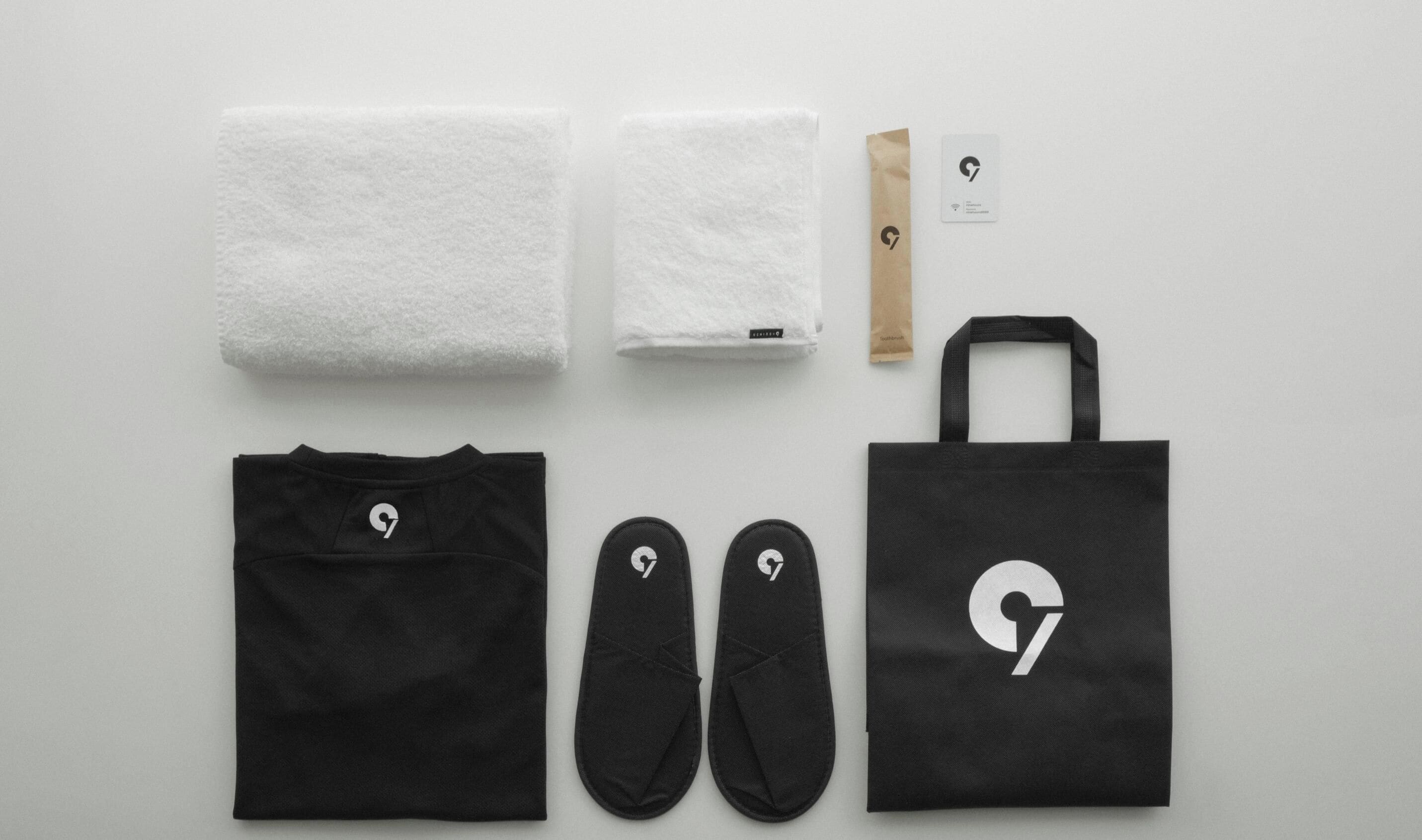 A black tote bag, a pair of black slippers, a black t-shirt, two white towels, a brown paper bag, and a card with a logo.