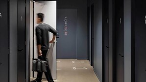 Un couloir moderne avec des flèches directionnelles peintes sur le sol, une personne portant un sac et des portes ornées de symboles indiquant les équipements disponibles.