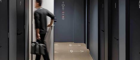 Un pasillo moderno con flechas direccionales en el suelo, una persona cargando un bolso y puertas con símbolos que indican servicios.