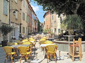 Un café al aire libre con sillas y mesas de mimbre amarillas, rodeado de edificios con balcones y plantas en macetas.
