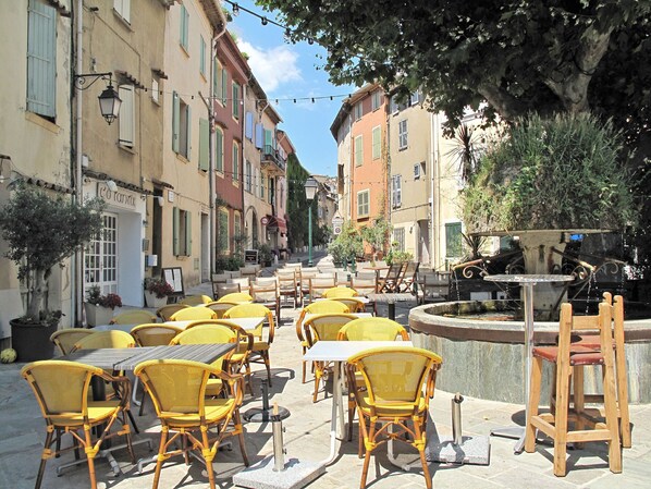 An outdoor café with yellow wicker chairs and tables, surrounded by buildings with balconies and potted plants.