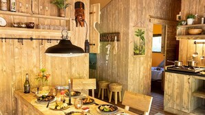A wooden dining area with a long table set for a meal, a hanging light fixture, and a rustic kitchen in the background. - Nice Tent Lodge Near a Lake (Mol)