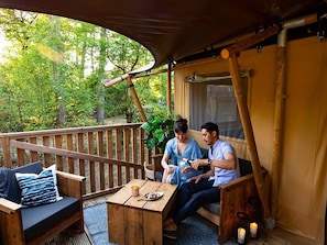 A wooden deck with a table and chairs, a covered area with a view of trees, and a window with curtains.