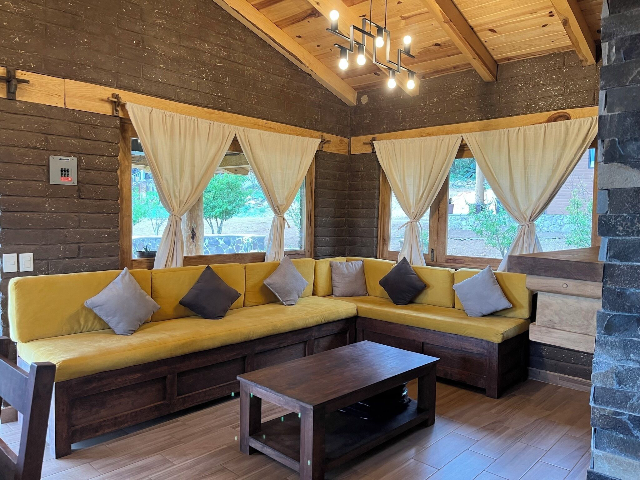 Una habitación acogedora con un sofá seccional amarillo, una mesa de centro de madera y ventanas amplias con cortinas.