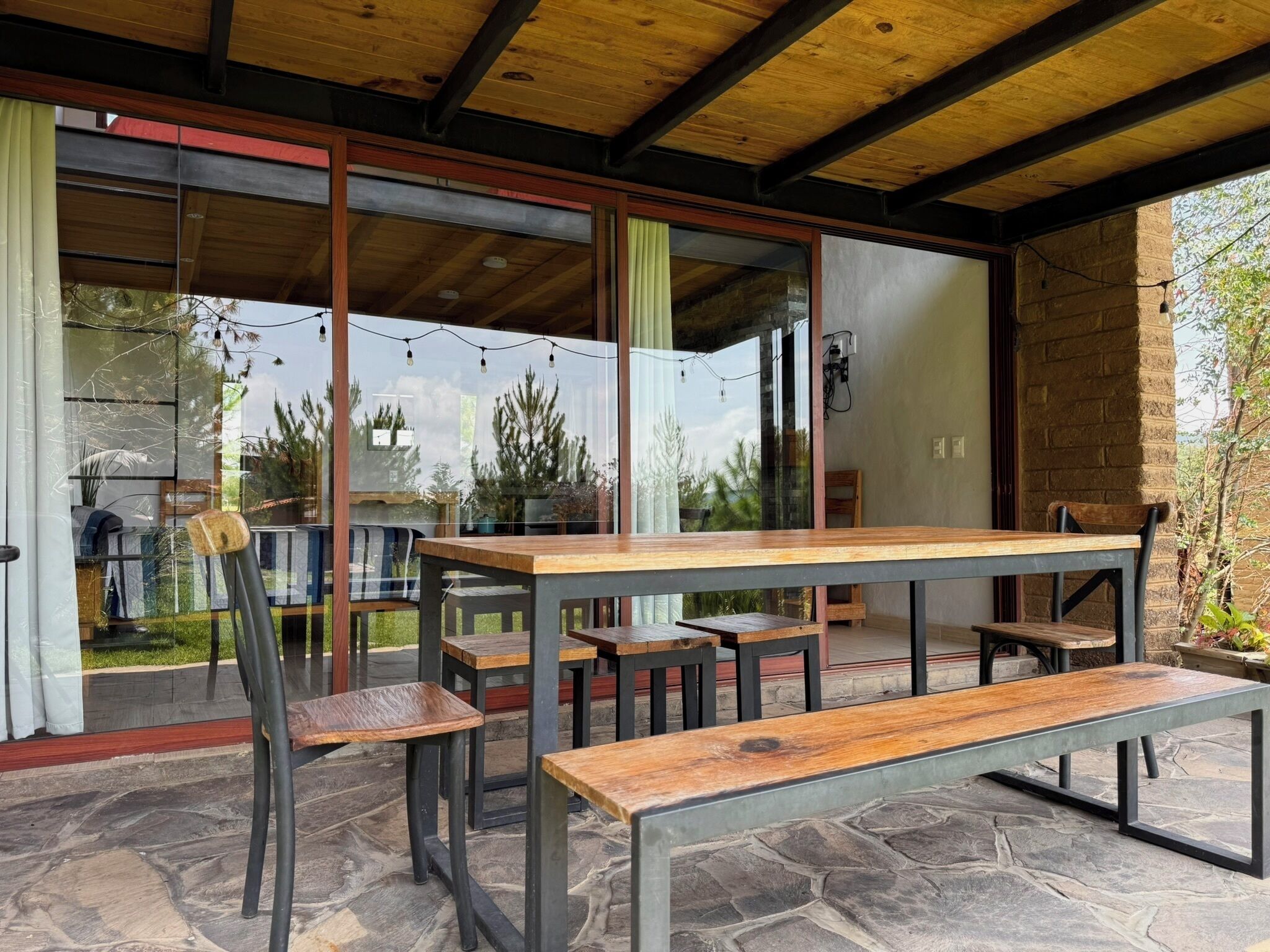 Un patio exterior con suelo de piedra, mesas y bancos de madera, y grandes puertas de cristal que dan a un jardín.