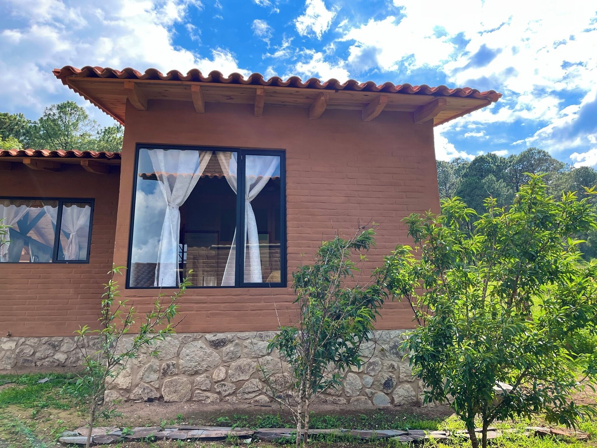 Una casa de dos pisos con cimientos de piedra, techo de tejas y un ventanal grande con cortinas blancas.