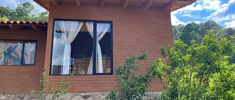 Una casa de dos pisos con cimientos de piedra, techo de tejas y un ventanal grande con cortinas blancas.