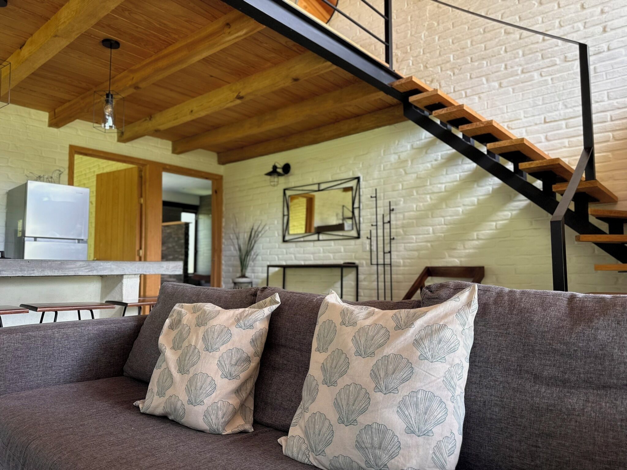 A modern living room with a wooden ceiling, a gray sofa with patterned cushions, a kitchen area in the background, and a staircase with a metal railing.