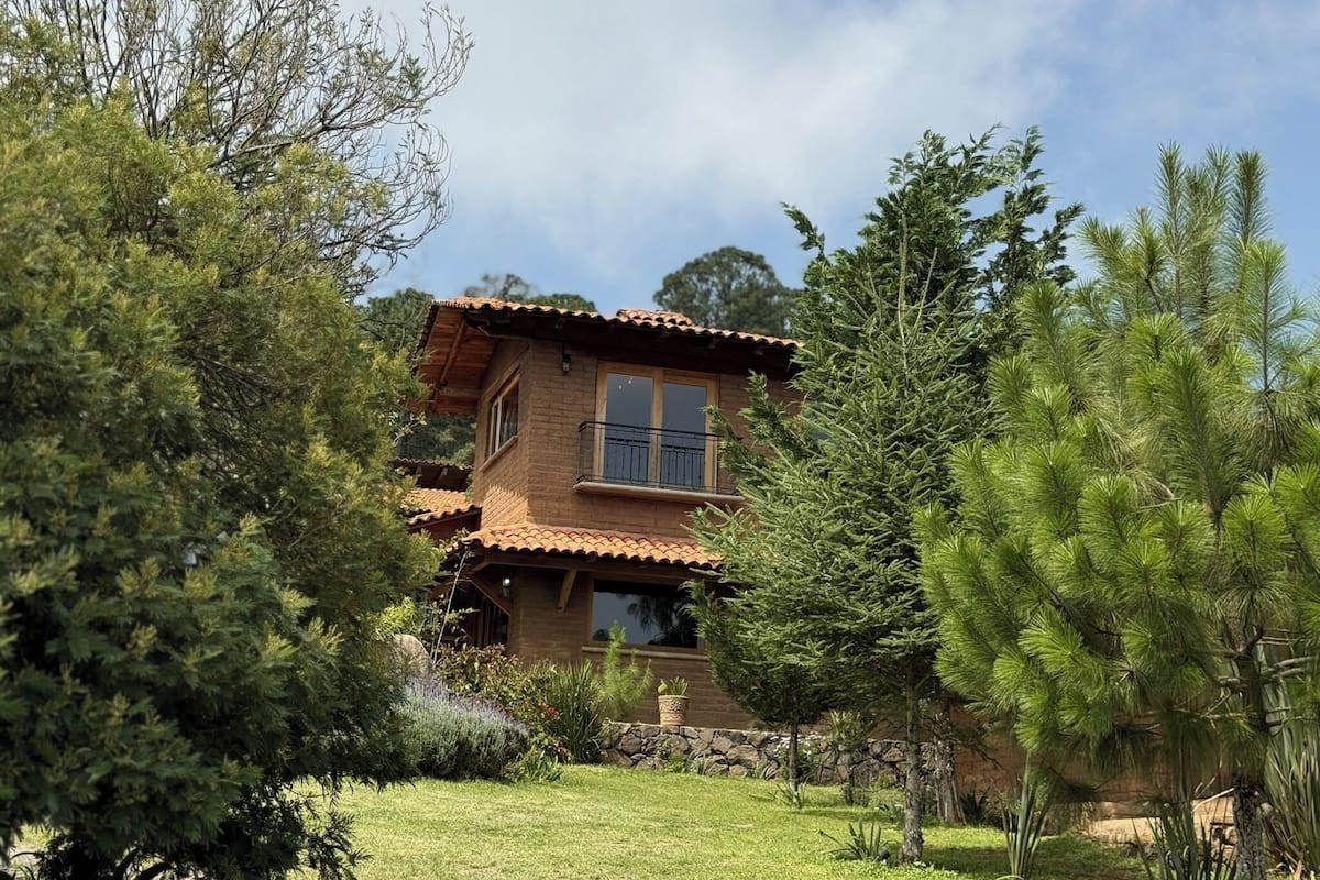 Une maison à deux étages, avec une façade en briques et un toit recouvert de tuiles, entourée d’une végétation luxuriante et d’arbres.