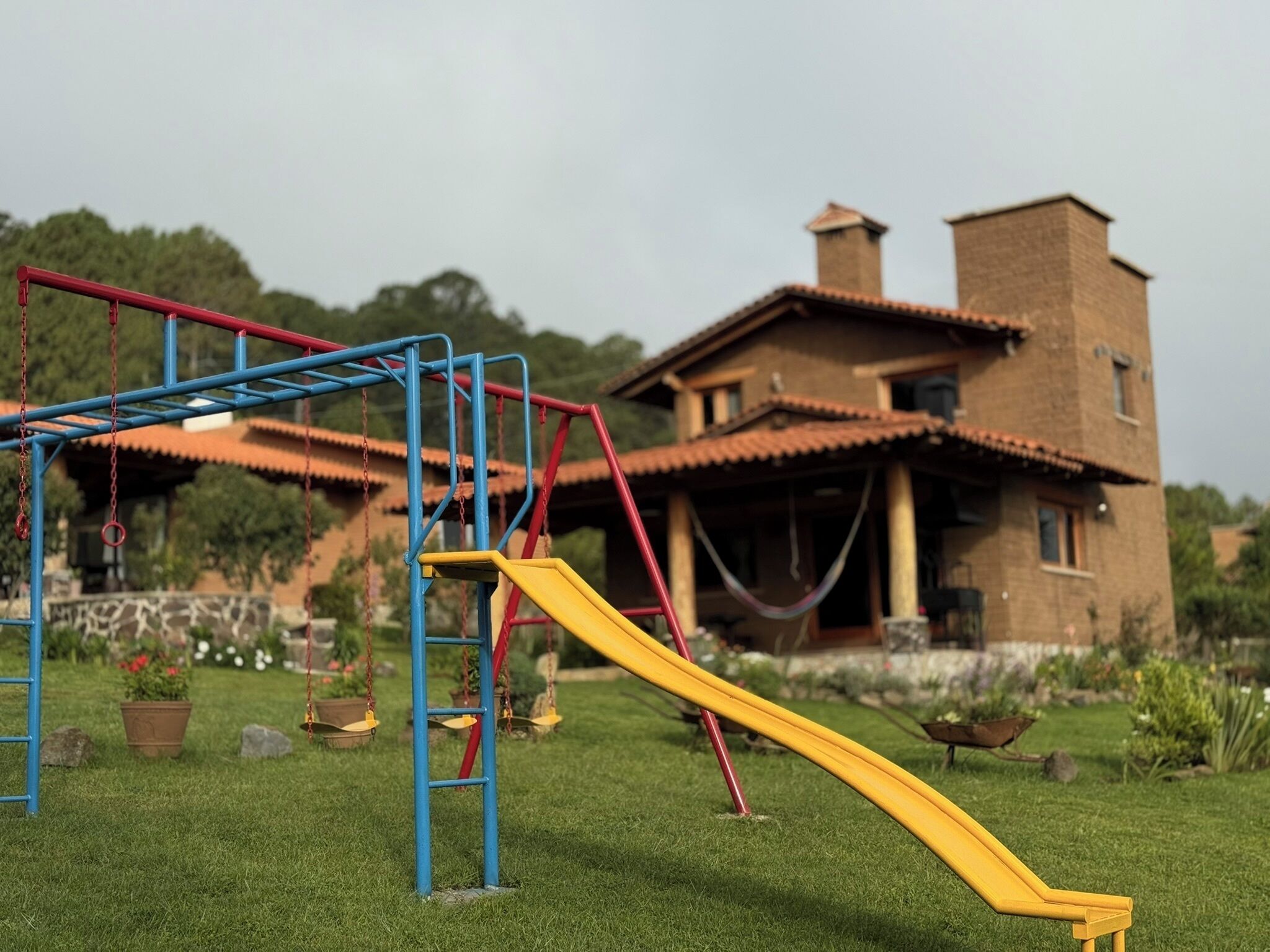 Un patio de juegos con un tobogán amarillo y estructuras azules y rojas, frente a una casa con techo de tejas.