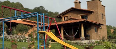 Un patio de juegos con un tobogán amarillo y estructuras azules y rojas, frente a una casa con techo de tejas.