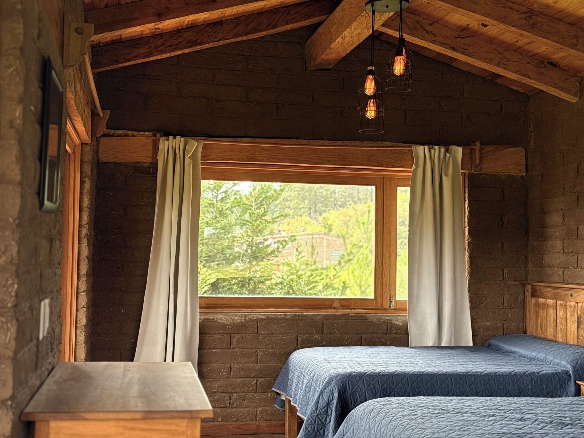 Una habitación rústica con dos camas, una ventana con cortinas, vigas de madera y lámparas colgantes.