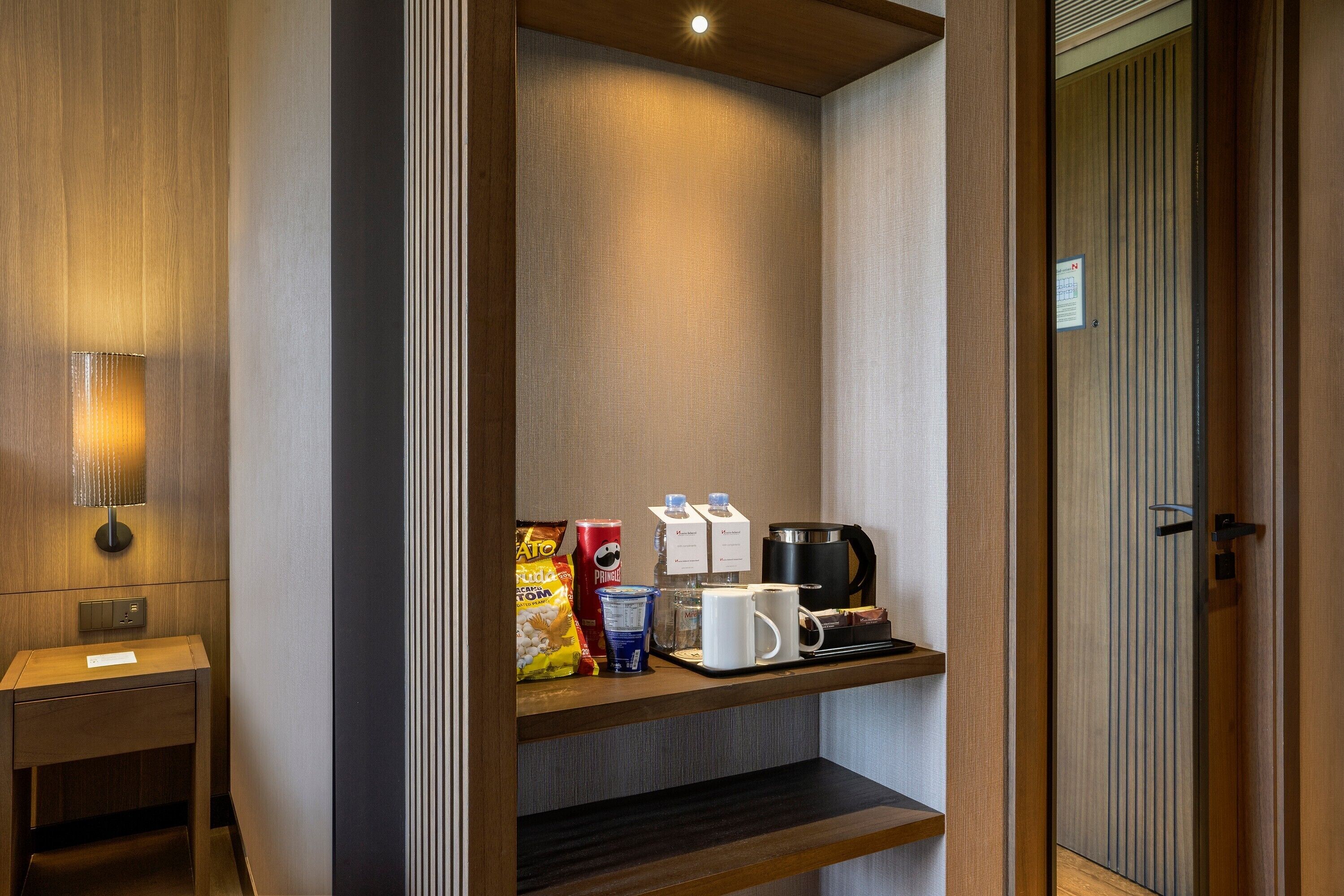 Habitación de hotel con mesita de noche de madera, lámpara de pared, repisa con cafetera, tetera, taza, cartón de leche, bolsa de papas y vaso de agua.