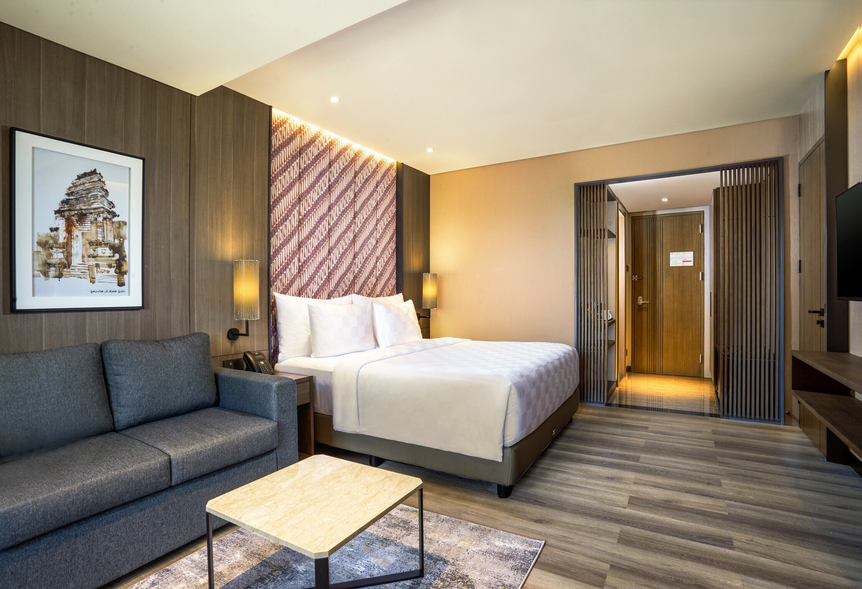 A modern hotel room with a bed, a sofa, a coffee table, and a wall-mounted TV.
