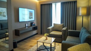 Una habitación de hotel moderna con un televisor de pantalla plana montado en la pared, un sofá cómodo, una mesita con una planta y un ventanal con cortinas.
