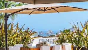 A wooden table with white chairs under a large umbrella, surrounded by tropical plants, with a view of the sea and mountains in the background. - Villa Cactus (Cabo San Lucas)