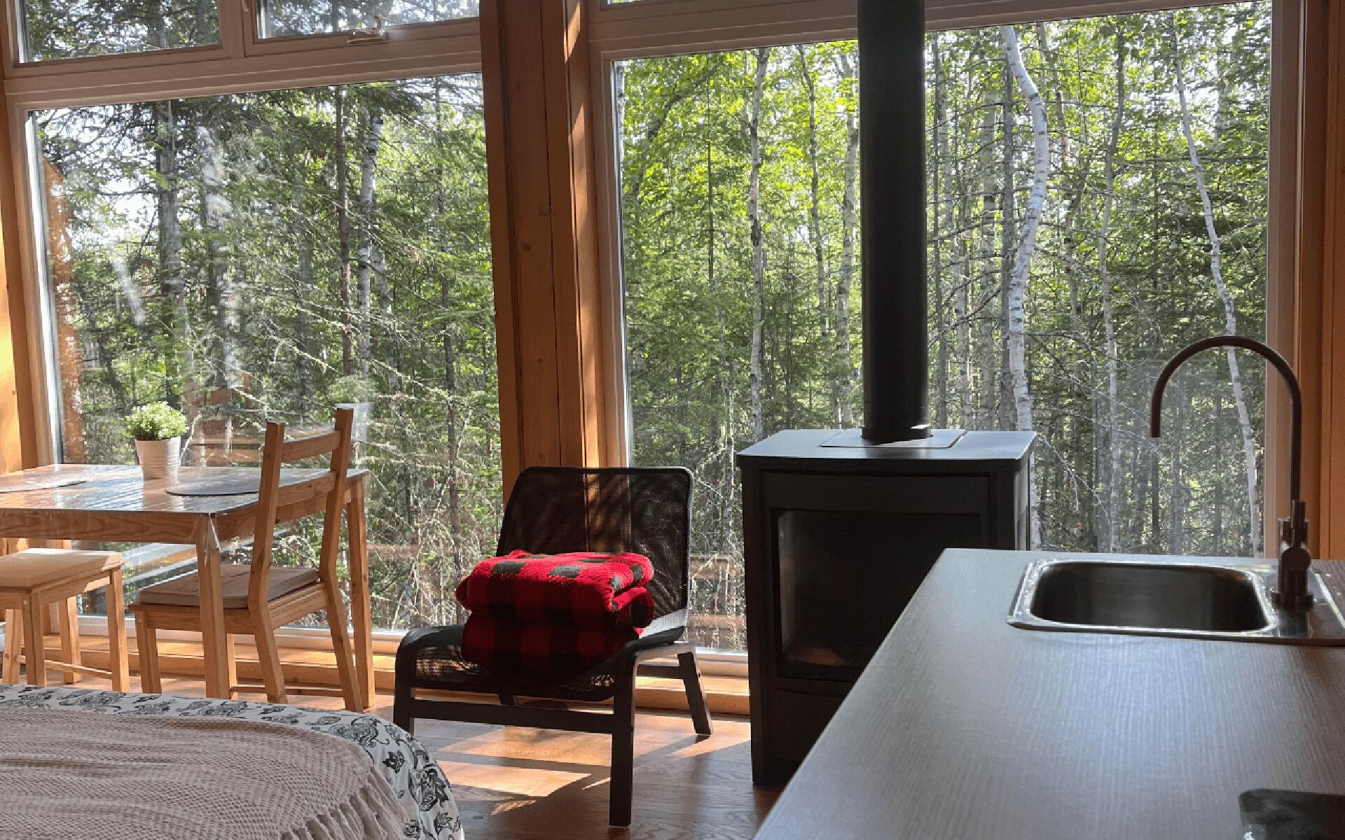 Interior acogedor de una cabaña con una mesa de comedor de madera, sillas, un sillón rojo a cuadros, un ventanal con vistas al bosque y una estufa de leña.
