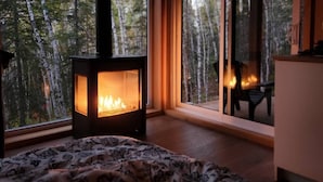 Una habitación acogedora con una estufa de leña, una cama con ropa de cama estampada y vistas a un bosque a través de amplias ventanas.