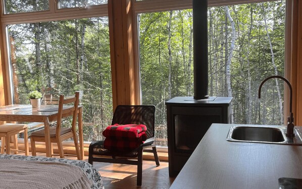 A cozy cabin interior with a wooden dining table, chairs, a red plaid chair, a large window with a forest view, and a wood-burning stove.