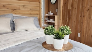 Una cama bien hecha con almohadas, una mesita de noche con una lámpara blanca y una estantería de madera con artículos de aseo.
