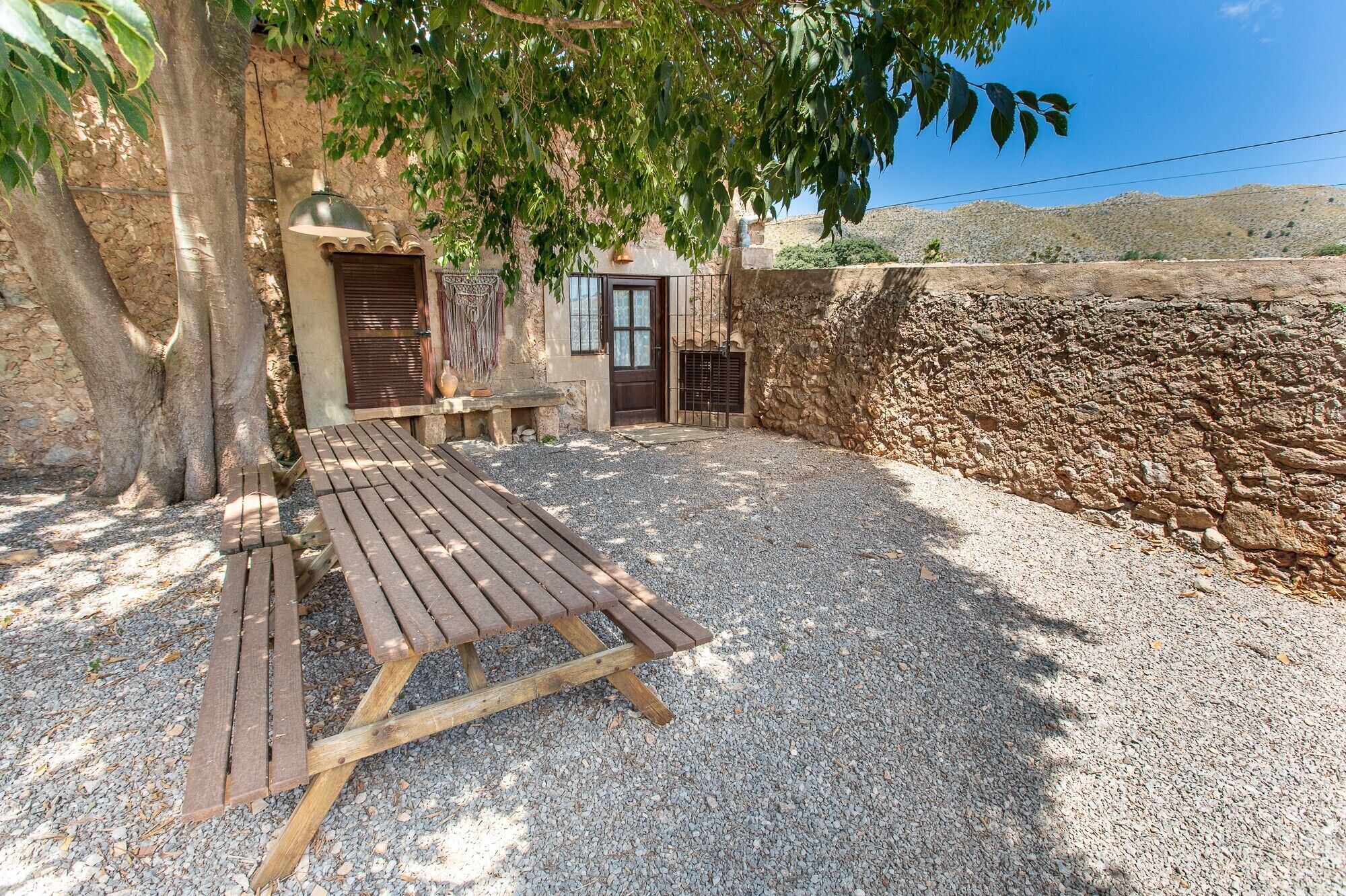 A wooden bench in a gravel area with a stone wall and a tree providing shade.