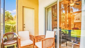 A balcony with wooden chairs and a plastic chair, a sliding glass door, and a view of a garden and a parked car.