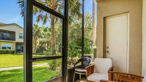 Un balcon donnant sur une pelouse verdoyante et des palmiers, une porte blanche et deux chaises.