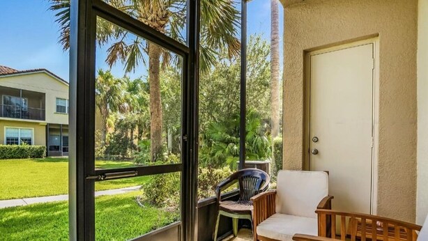 A balcony with a view of a green lawn and palm trees, a white door, and two chairs.
